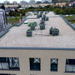 Dachowe Love - Nowoczesny, płaski dach budynku wielorodzinnego pokryty żwirem, z widocznymi elementami wentylacji i obróbkami blacharskimi. Ujęcie z góry, w tle zabudowa miejska.