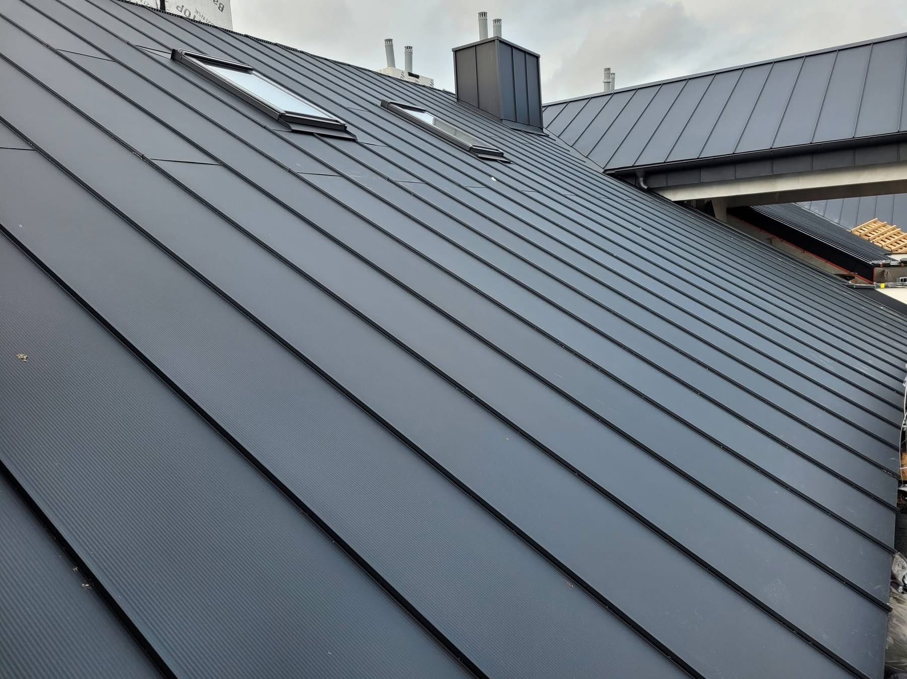 Nowoczesny, grafitowy dach z blachy na rąbek stojący, z oknami dachowymi i kominem. Widok z dołu ukazuje precyzję wykonania i regularność linii.