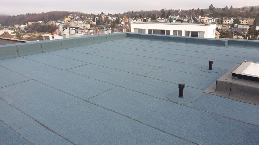 Płaski dach pokryty papą termozgrzewalną z widocznymi kominkami wentylacyjnymi i świetlikiem. W tle zabudowa miejska w pochmurny dzień.