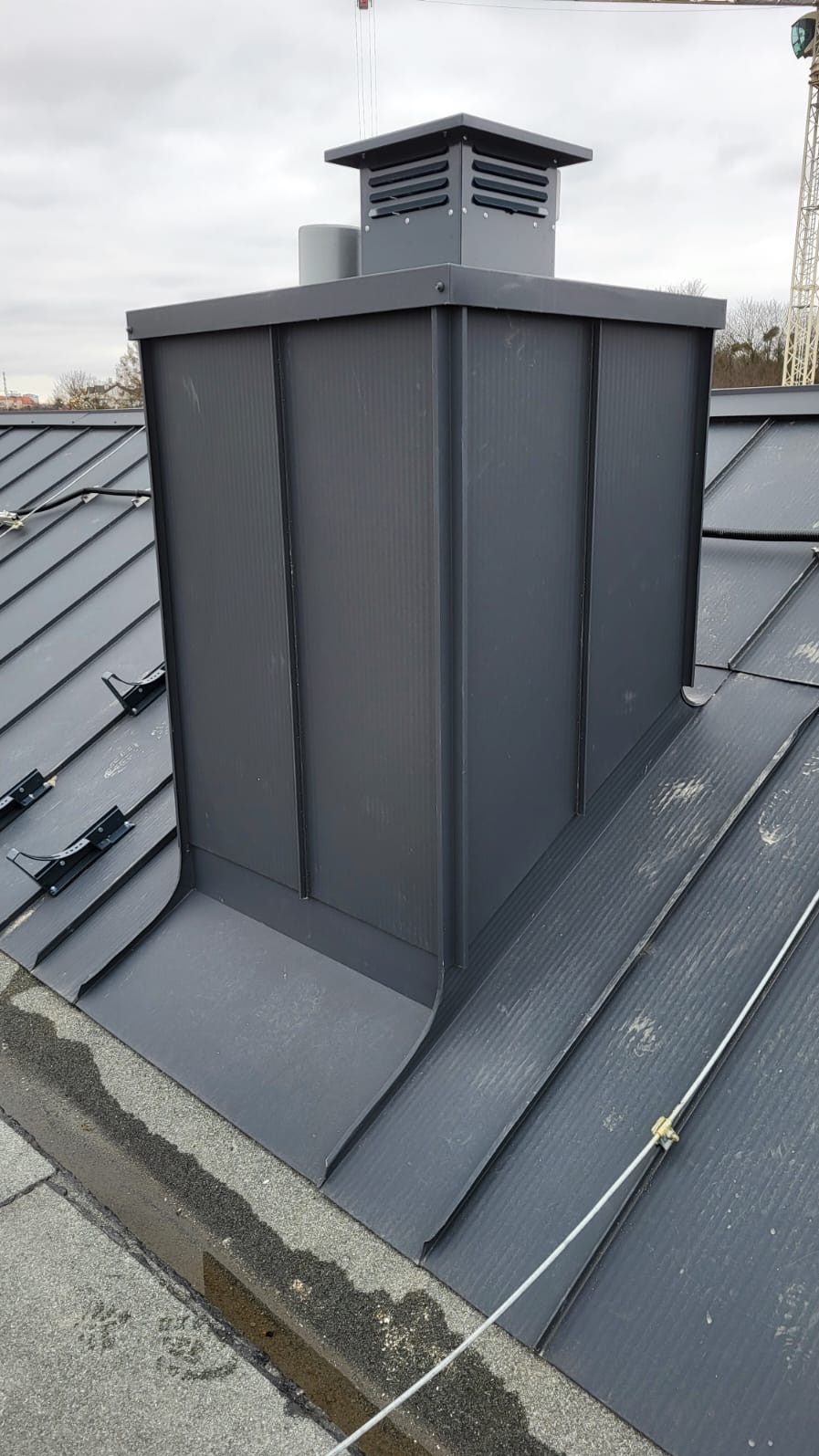 Szary komin wentylacyjny na dachu z blachy trapezowej, z elementami mocującymi. Widoczne łączenia blach i detale wykończeniowe. W tle fragment konstrukcji budowlanej.