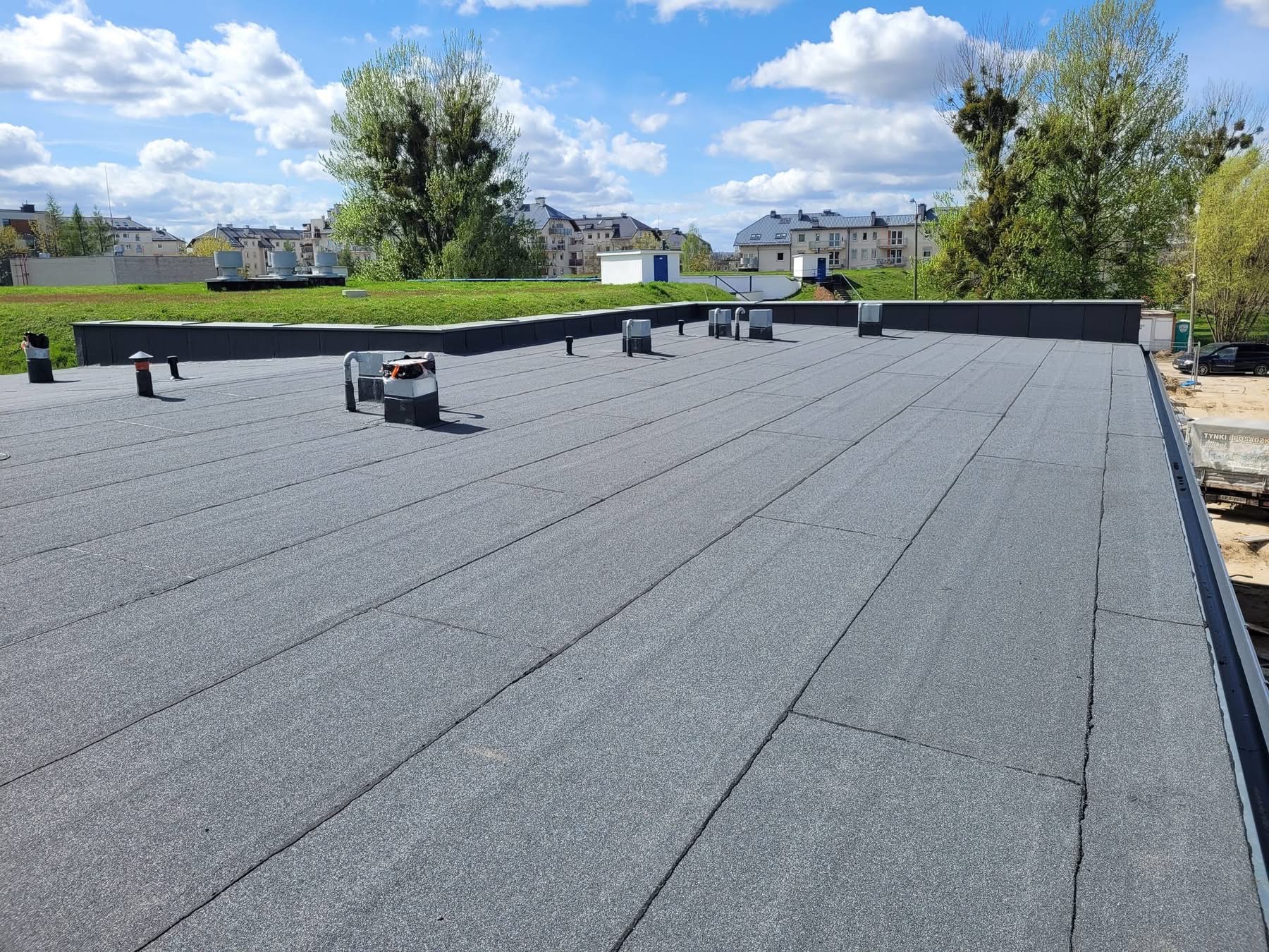 Szary, płaski dach pokryty papą termozgrzewalną z elementami wentylacyjnymi i zielenią w tle. Widoczne budynki mieszkalne i błękitne niebo.