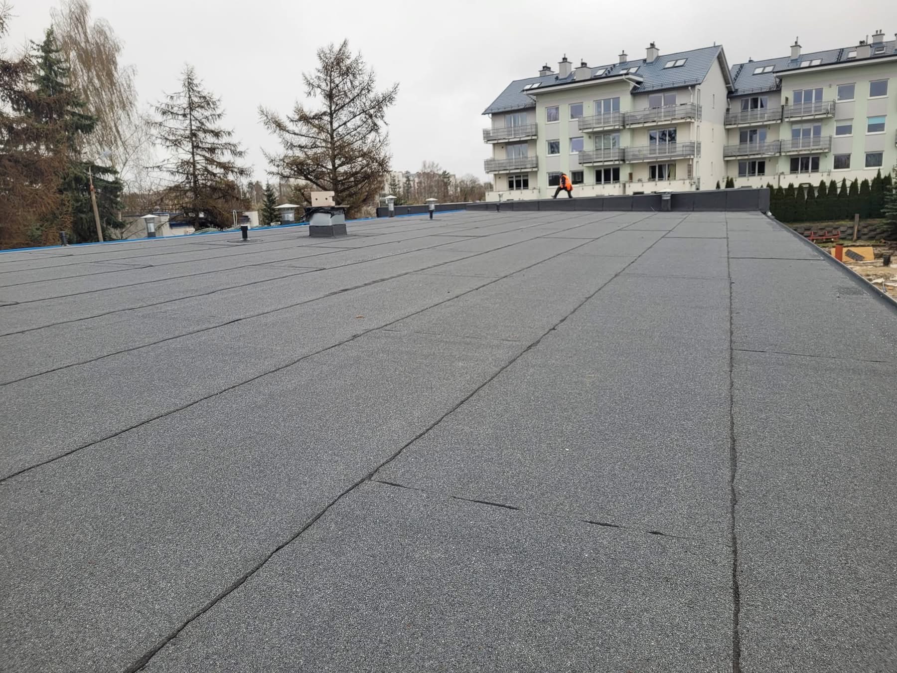 Wykończony płaski dach pokryty papą termozgrzewalną na budynku wielorodzinnym. Widoczna osoba w odblaskowej kamizelce na krawędzi dachu.