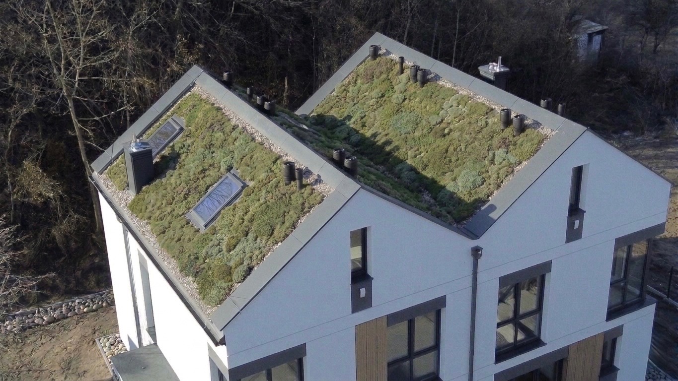 Nowoczesny dom z dachem pokrytym zielenią, widok z góry. Dach z oknami dachowymi i systemem odprowadzania wody. Biała elewacja budynku z ciemnymi obramowaniami okien.