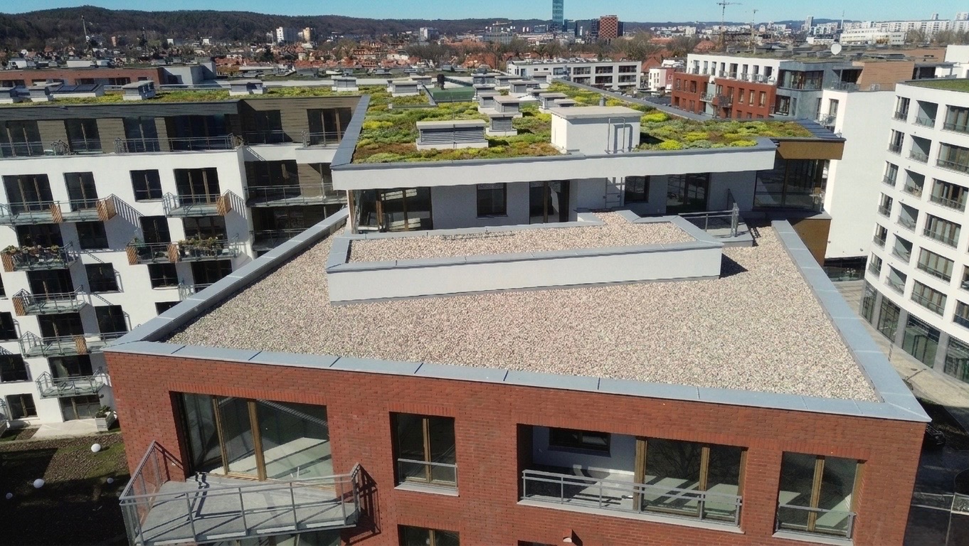 Nowoczesny budynek z płaskim dachem pokrytym drobnym żwirem i zielenią. Widok z góry na architekturę miejską w słoneczny dzień.