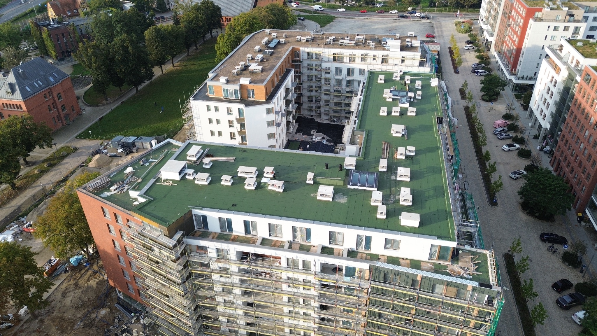 Zielony dach nowego budynku wielorodzinnego z licznymi wywietrznikami, widziany z góry. Na elewacji rusztowania. W tle inne budynki i zieleń miejska.