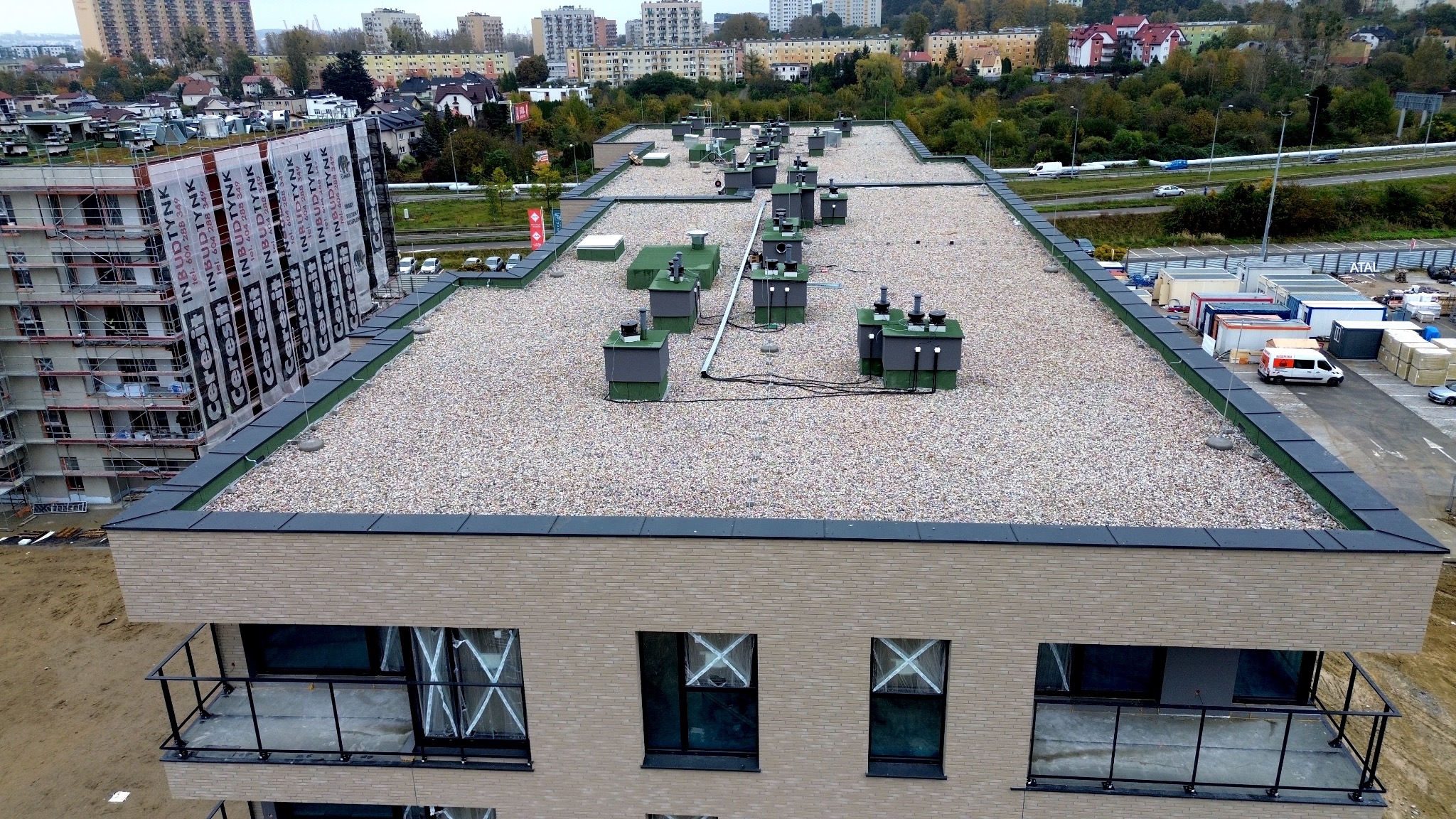 Nowoczesny, płaski dach budynku wielorodzinnego pokryty żwirem, z widocznymi elementami wentylacji i obróbkami blacharskimi. Ujęcie z góry, w tle zabudowa miejska.