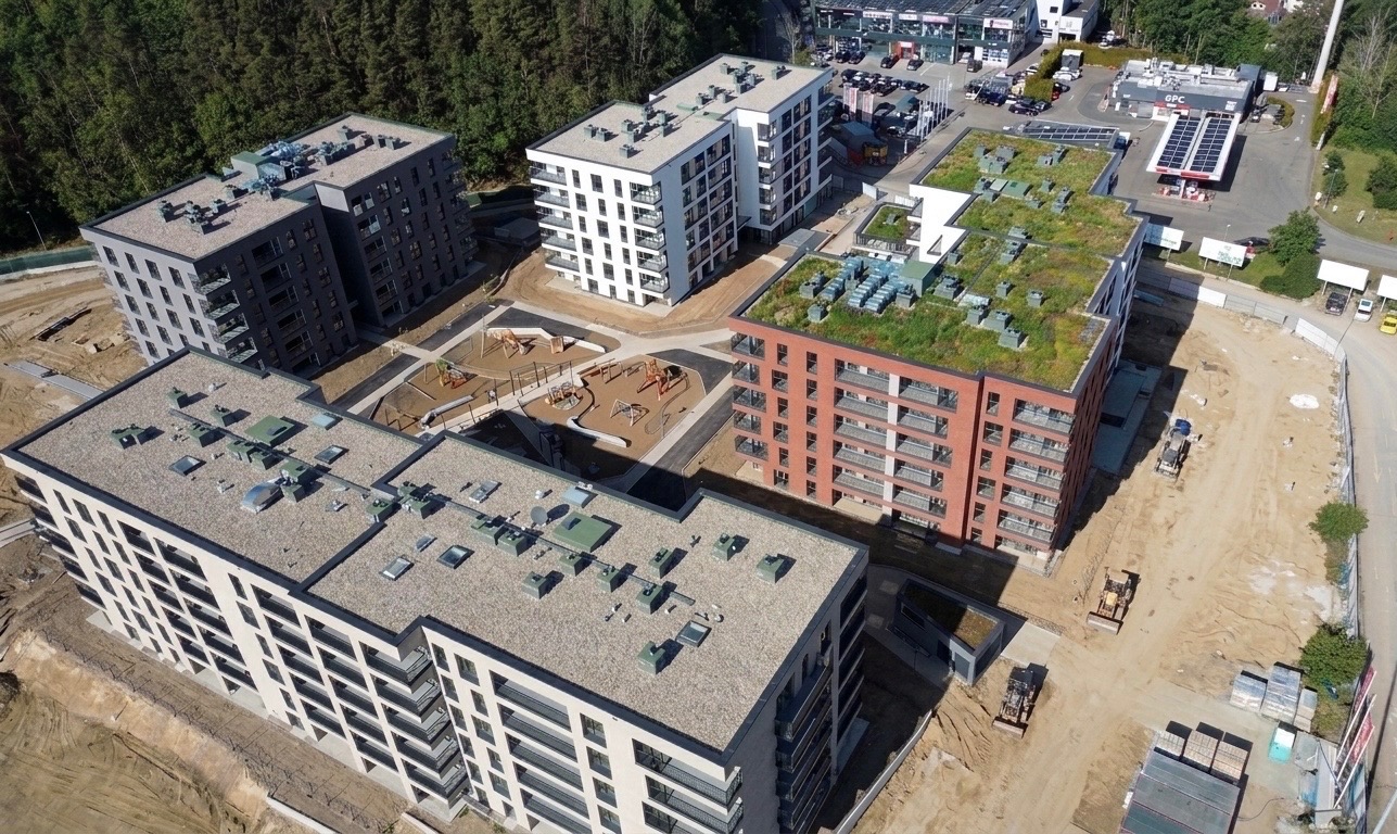 Widok z lotu ptaka na kompleks budynków mieszkalnych z dachami pokrytymi zielenią, placem zabaw i terenem w trakcie zagospodarowywania. Ujęcie z góry.