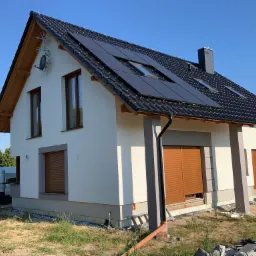 Nowoczesny dom jednorodzinny z panelami fotowoltaicznymi na dachu, jasną elewacją i brązowymi roletami zewnętrznymi, widok z zewnątrz, w tle biały bus i trawa.