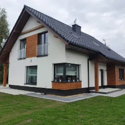 Nowoczesny dom jednorodzinny z białą elewacją, drewnianymi akcentami i czarnym dachem, widok z zewnątrz na świeżo położony trawnik i betonowy chodnik.