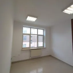 Puste, jasne pomieszczenie po remoncie z białymi ścianami, oknem z widokiem na budynek i dwoma lampami sufitowymi.