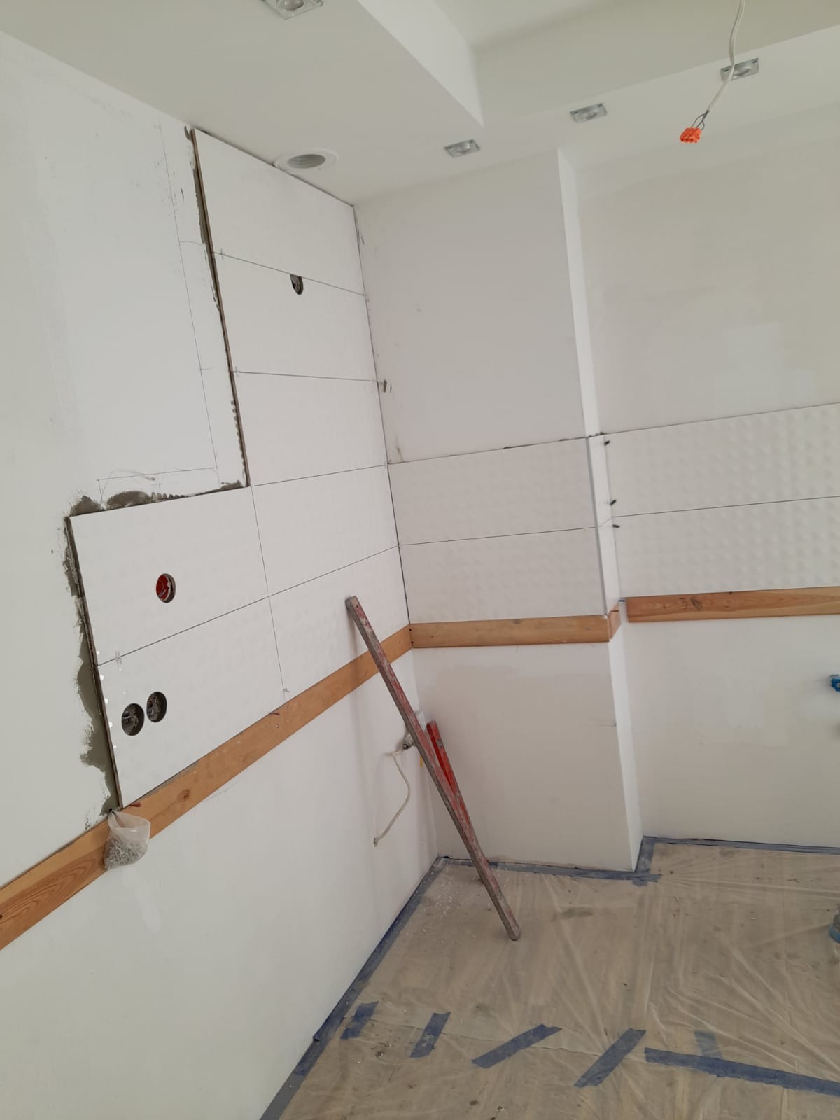 Białe płytki z subtelnym wzorem układane na ścianie w kuchni, widoczne otwory na gniazdka elektryczne, drewniane listwy pomocnicze i poziomnica oparta o ścianę, folia ochronna na podłodze.