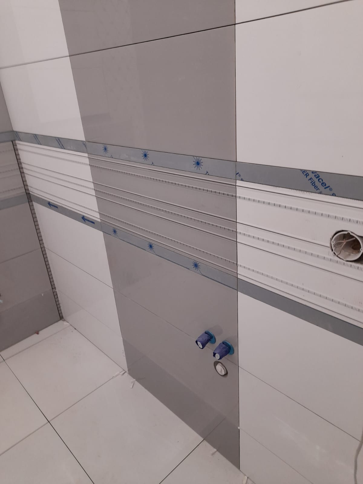 Świeżo wykafelkowana ściana w łazience z widocznymi przyłączami wodnymi zabezpieczonymi niebieską folią, szaro-białe płytki z dekoracyjnym paskiem.