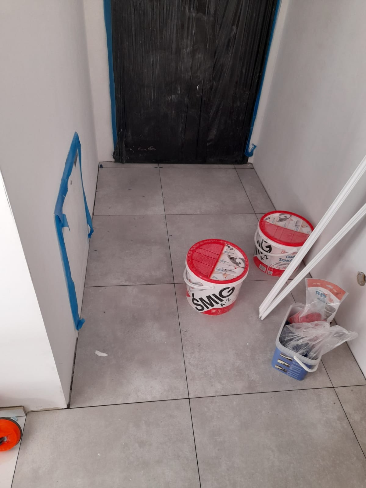 Przygotowane pomieszczenie do prac wykończeniowych: podłoga wyłożona szarymi płytkami, ściany zabezpieczone taśmą malarską, zakryte folią drzwi, widoczne dwa wiadra z masą szpachlową oraz narzędzia.