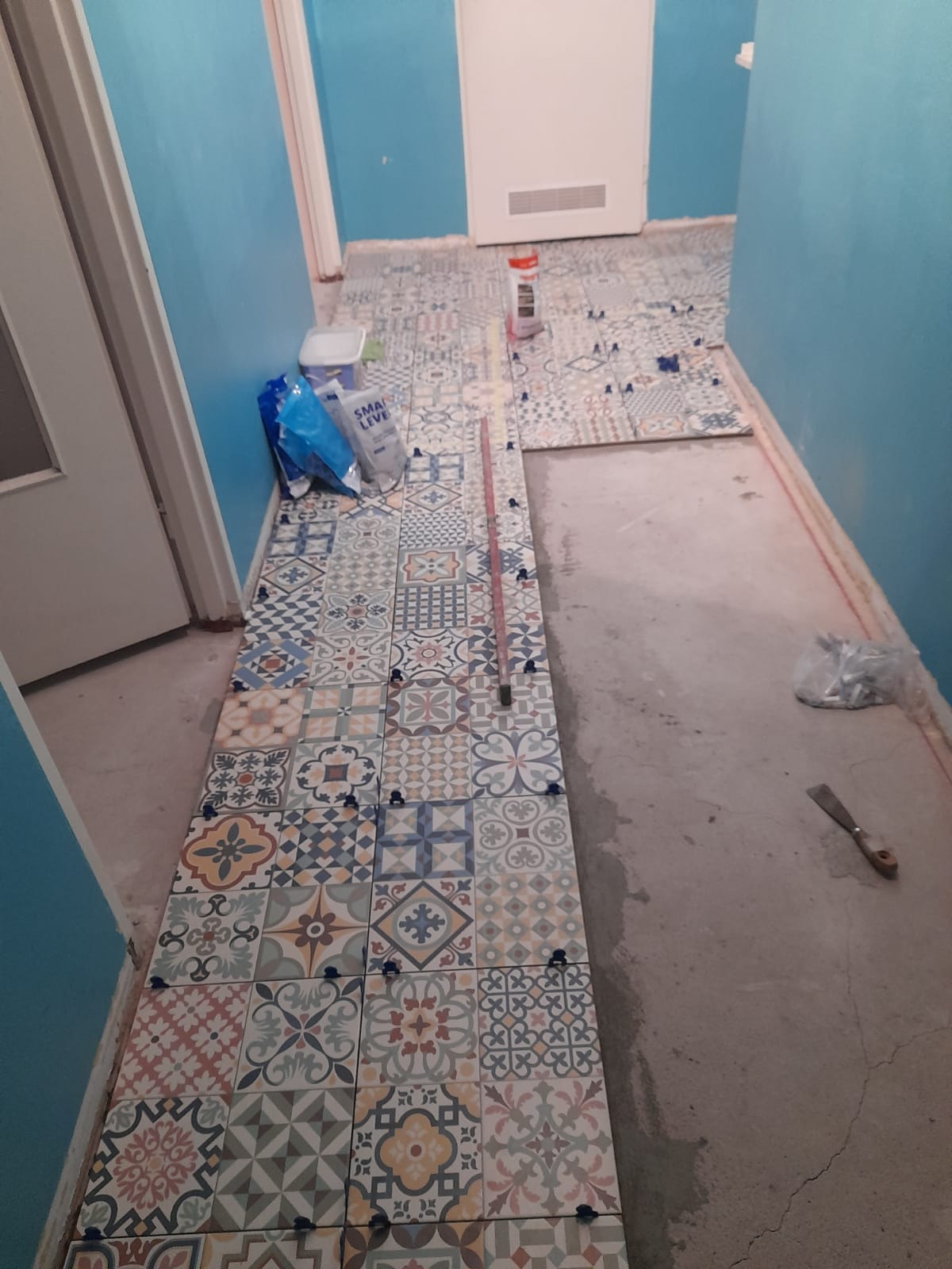 Układanie wzorzystych płytek podłogowych w korytarzu, widoczne krzyżyki dystansowe, paczka z klejem, poziomica i szpachelka.