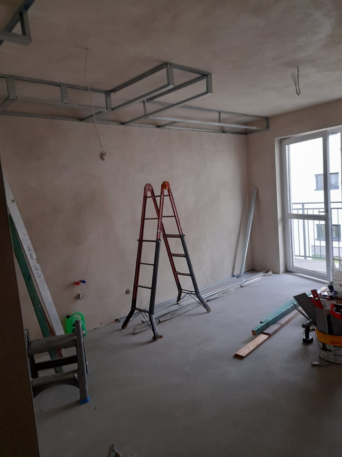 Pokój w trakcie remontu: surowe ściany po szpachlowaniu, metalowa konstrukcja sufitu podwieszanego, drabina, profile i narzędzia budowlane.