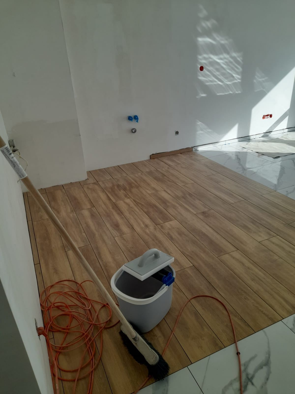 Fragment pokoju z nowo ułożonymi panelami podłogowymi imitującymi drewno, widoczny mop, wiadro z wyciskaczem i pomarańczowy przedłużacz przy ścianie.