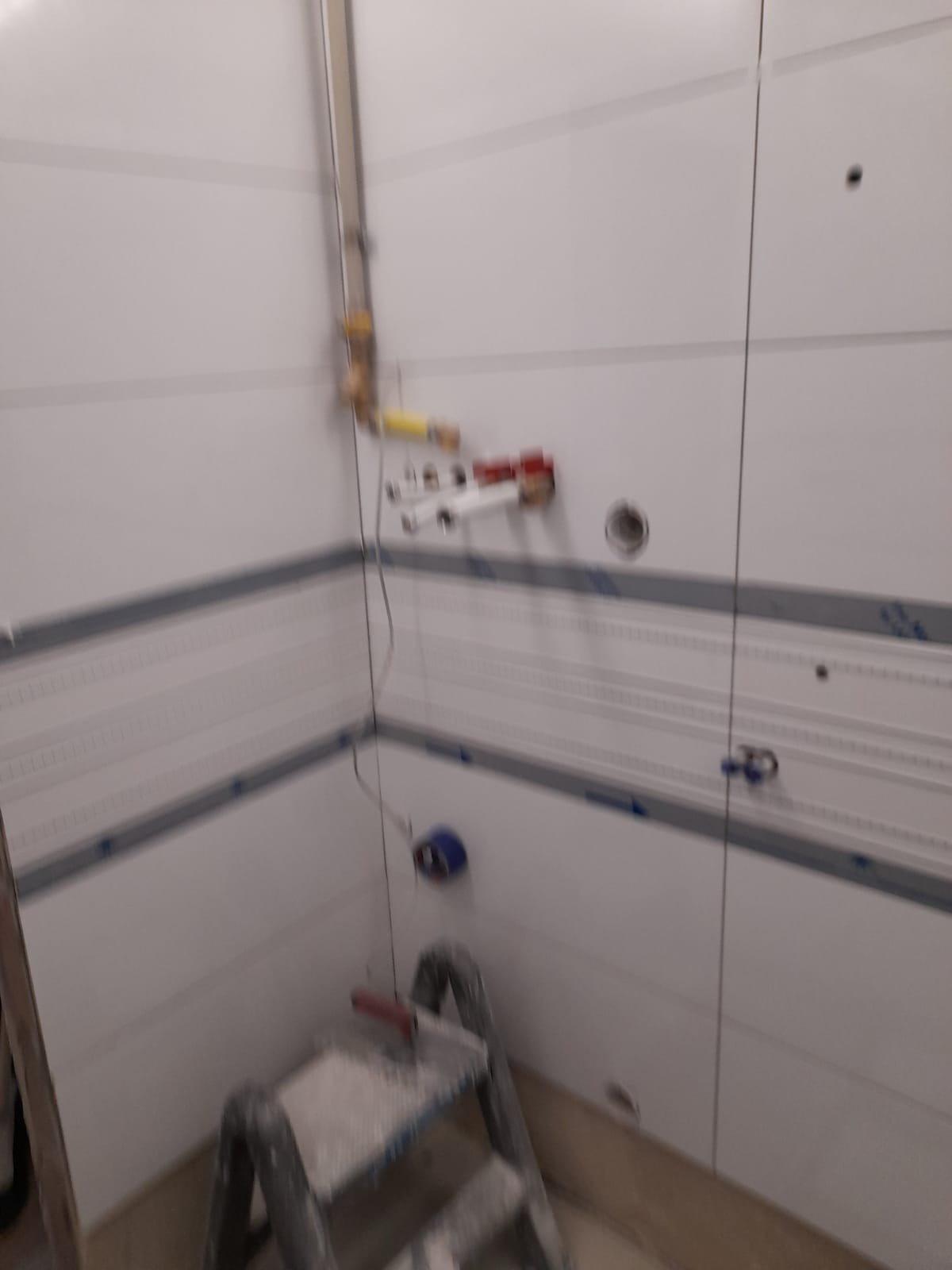 Ściana w trakcie wykańczania płytkami z widoczną instalacją hydrauliczną i drabinką malarską, przygotowana do dalszych prac.