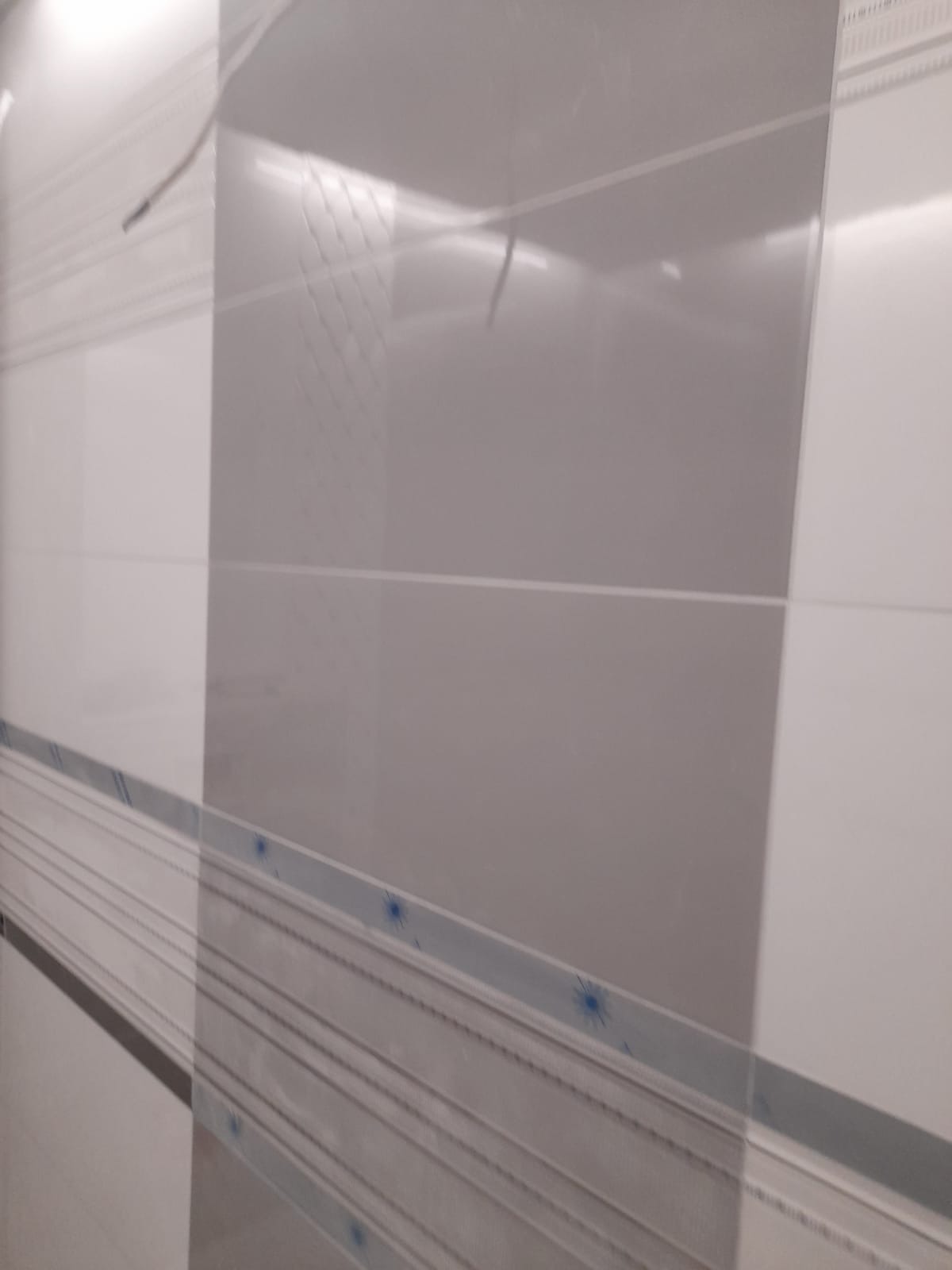 Biało-szare, błyszczące płytki ścienne z dekoracyjnym, poziomym pasem z niebieskimi akcentami, ułożone w regularny wzór.