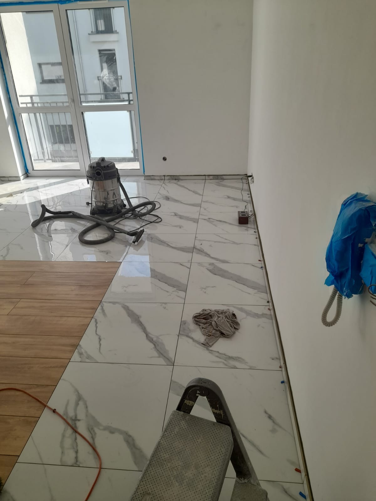Wnętrze pokoju w trakcie wykańczania podłogi: płytki imitujące marmur przechodzą w panele podłogowe, widoczny odkurzacz przemysłowy i drabina.