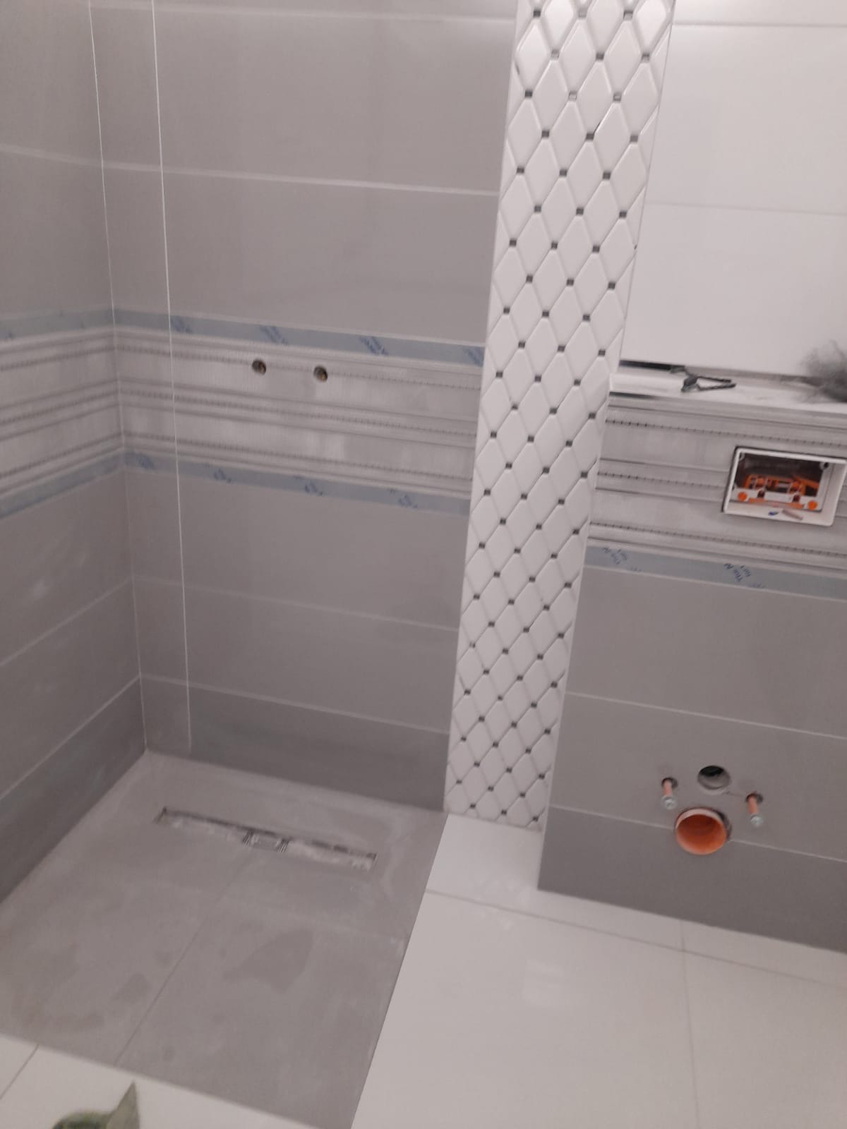 Wnętrze łazienki w trakcie remontu: szare płytki ścienne z dekoracyjnym pasem, pionowy panel z białych płytek w romby, widoczny odpływ liniowy w podłodze i instalacja sanitarna.