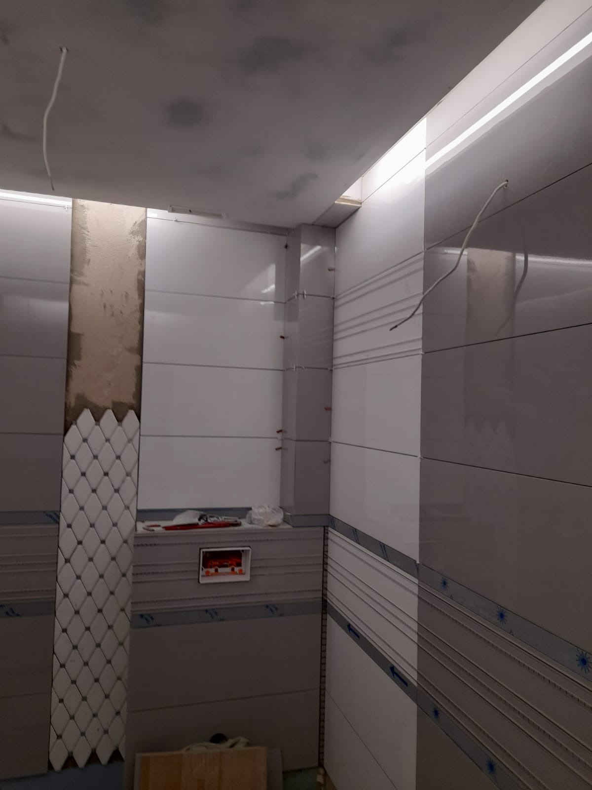 Wnętrze łazienki w trakcie prac wykończeniowych: częściowo ułożone białe płytki z dekoracyjnym pasem i mozaiką, widoczne narzędzia, wystające przewody elektryczne.