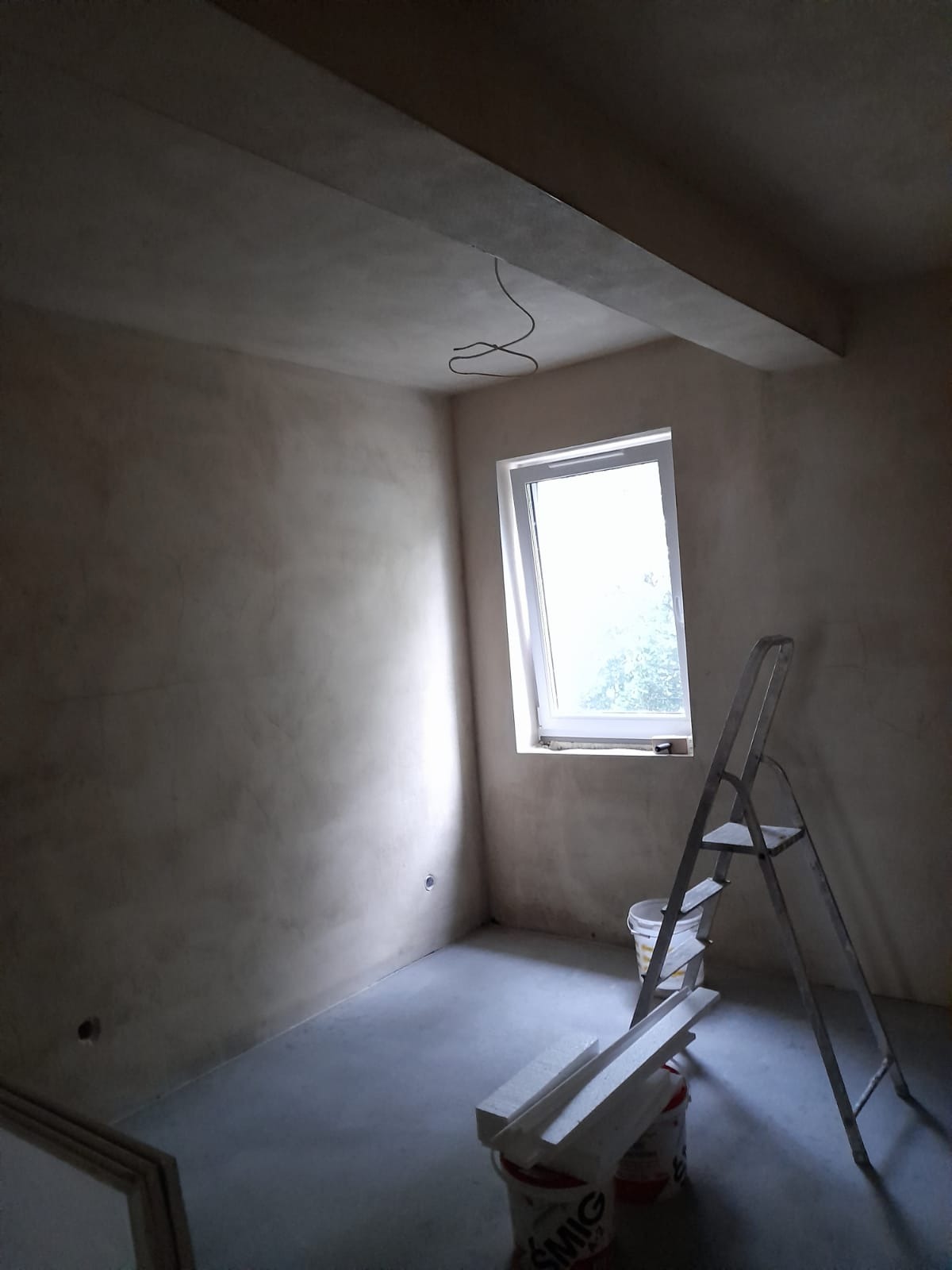 Pokój w trakcie wykańczania: ściany pokryte szpachlą, widoczna drabina, wiadro z materiałami budowlanymi i styropianowe listwy przypodłogowe.