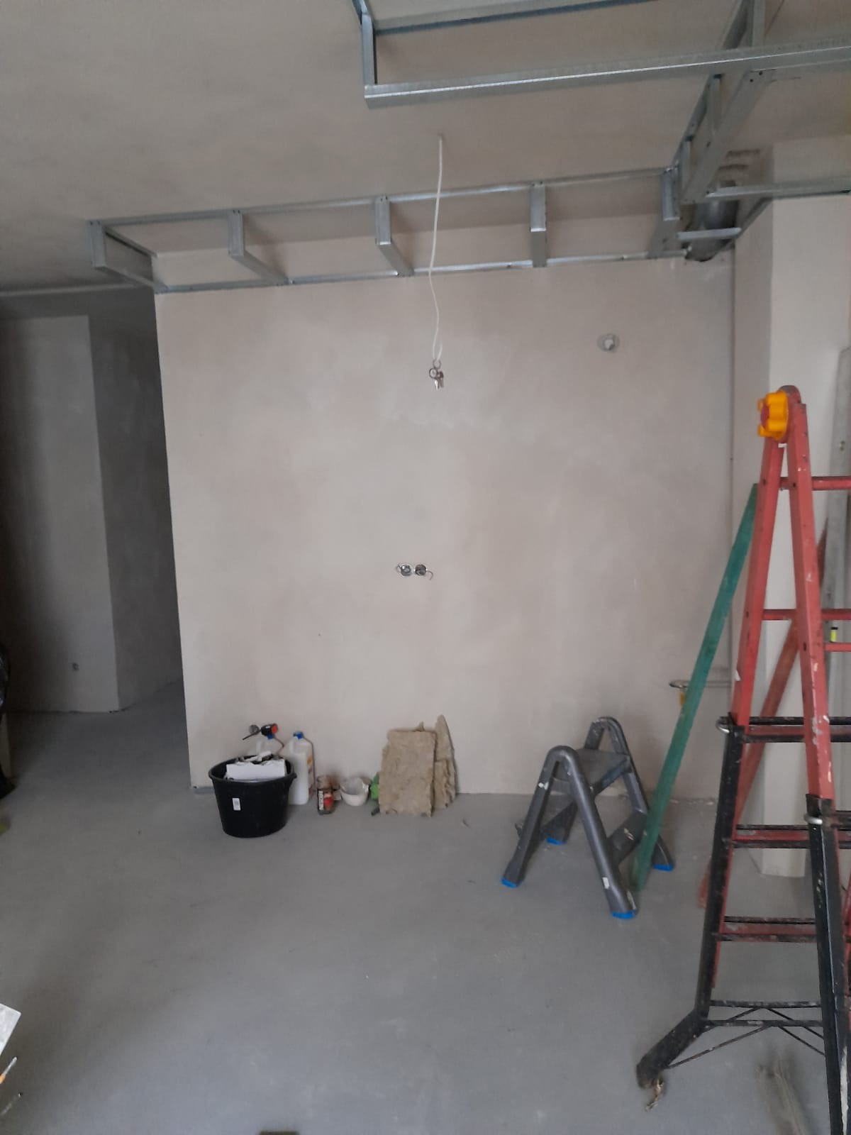 Przygotowanie do prac wykończeniowych: ściana po szpachlowaniu, widoczne profile sufitowe, drabina, wiadro z narzędziami i materiałami budowlanymi.