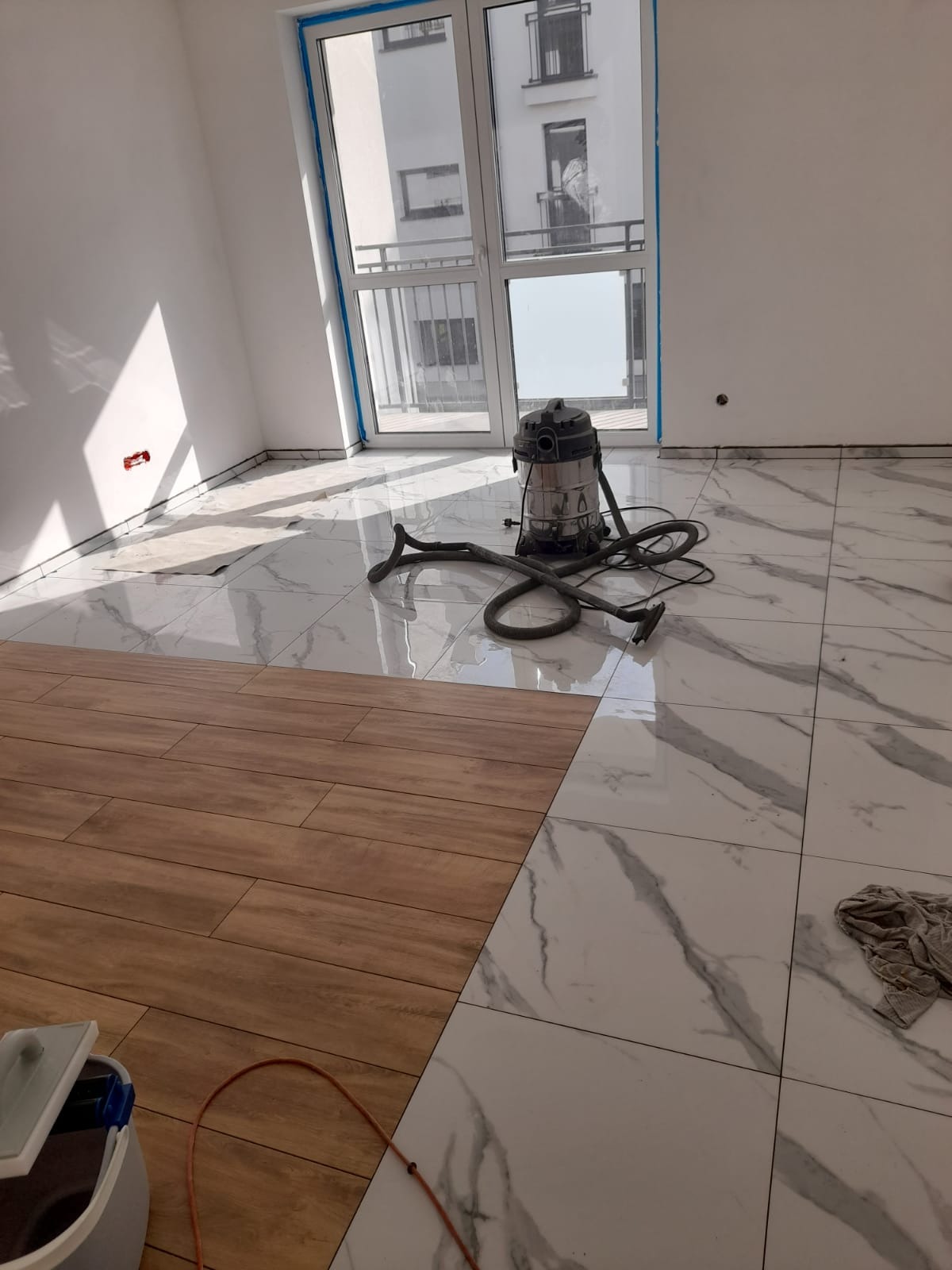 Układanie parkietu w pokoju z marmurowymi płytkami, widoczny odkurzacz przemysłowy, fragment okna balkonowego i wiadro z klejem