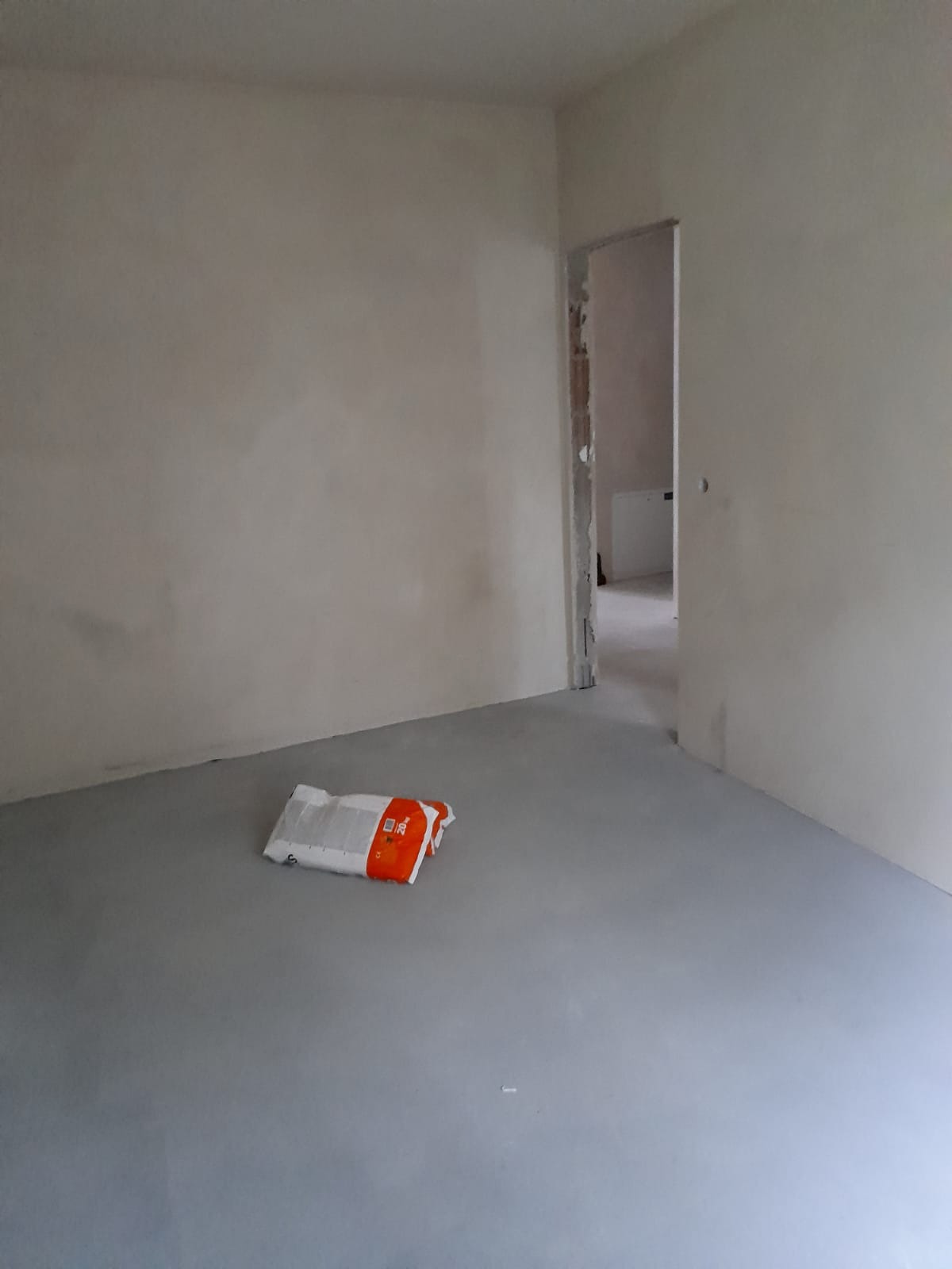 Pokój w trakcie remontu: ściany po szpachlowaniu, gładki, szary podkład podłogowy, worek z zaprawą na podłodze, otwarte drzwi do sąsiedniego pomieszczenia.