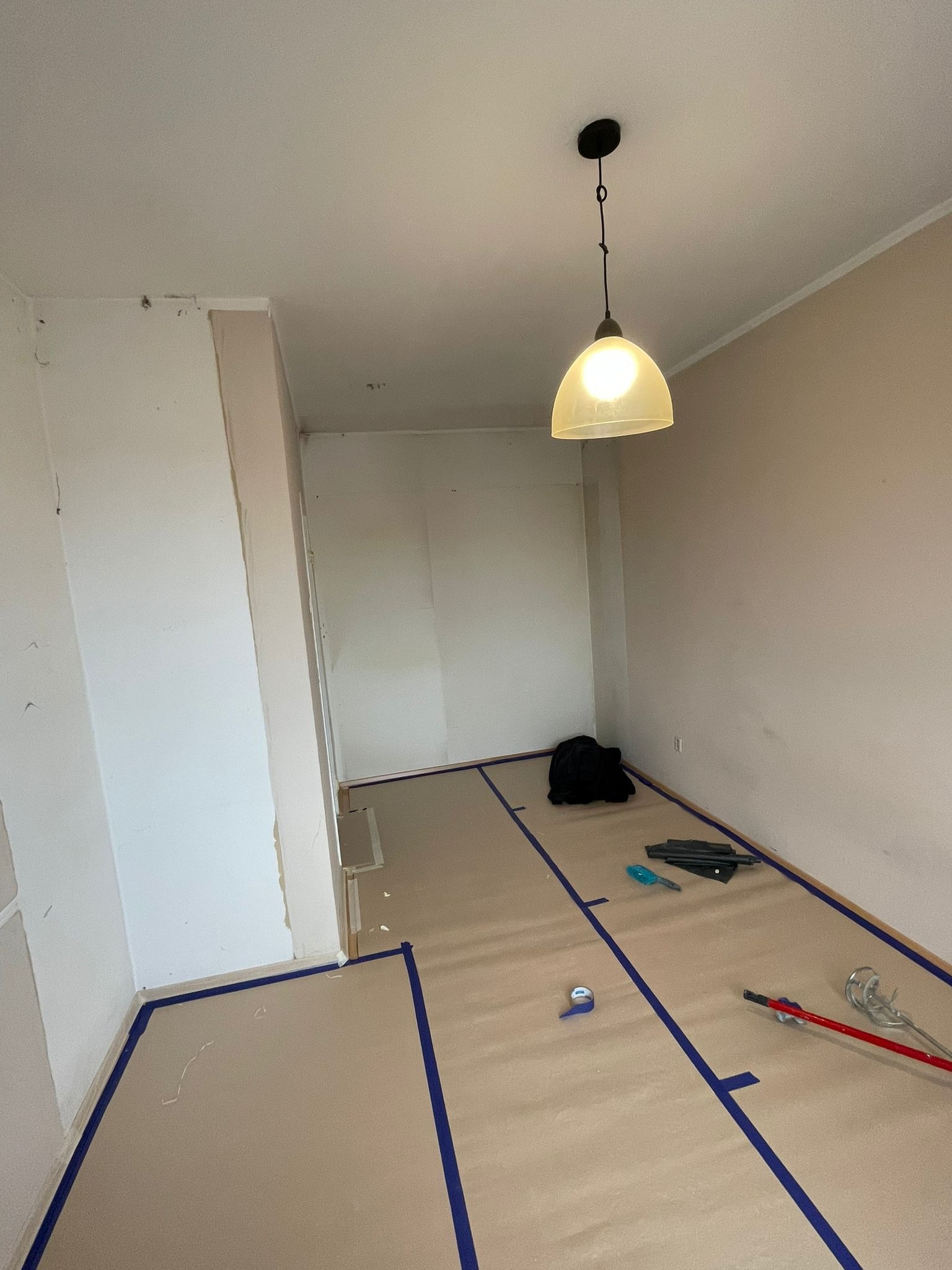 Przygotowane pomieszczenie do malowania: podłoga zabezpieczona papierem i taśmą malarską, widoczne narzędzia malarskie i lampa wisząca.