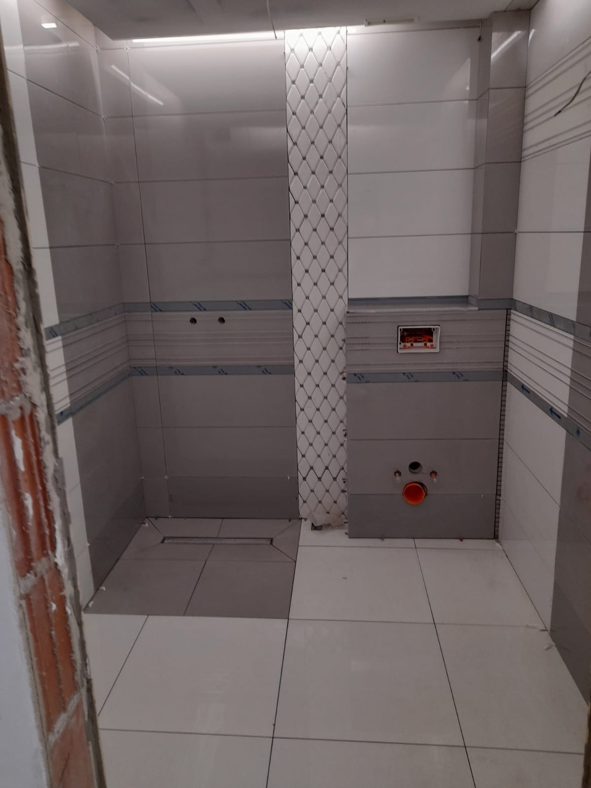 Wnętrze łazienki w trakcie remontu, widoczne ułożone płytki ścienne w odcieniach szarości i bieli, fragment niezabezpieczonego ceglanego muru, oraz instalacja sanitarna z pomarańczową rurą.