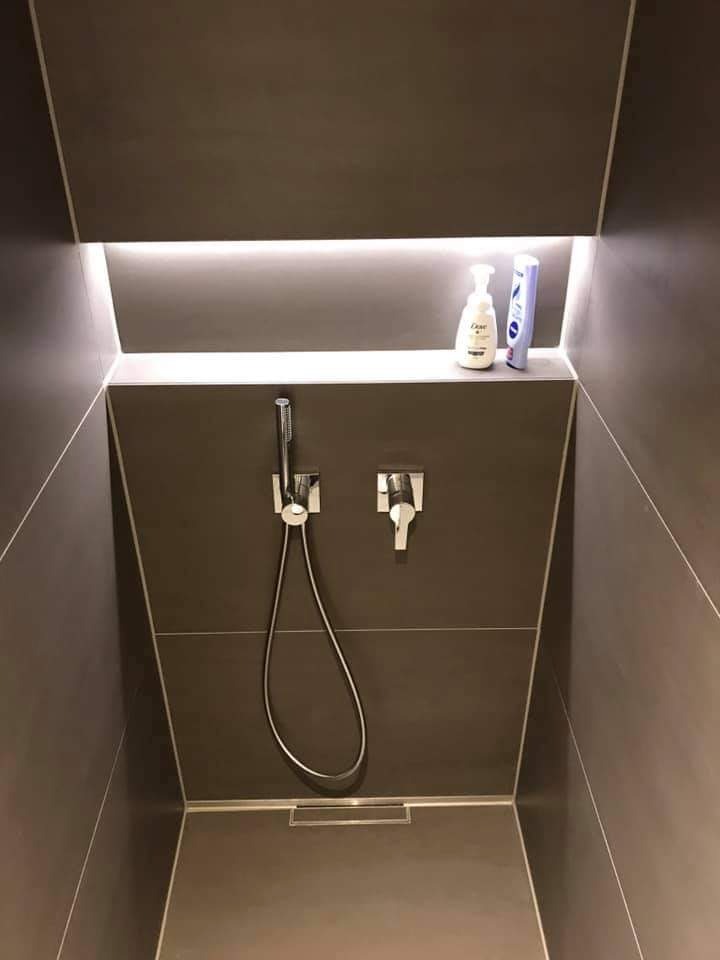 Nowoczesny prysznic z szarymi płytkami, wbudowanym oświetleniem LED nad półką z kosmetykami, minimalistyczna armatura.