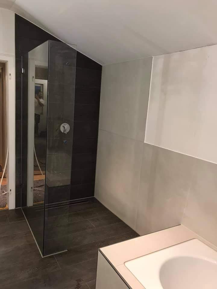 Nowoczesna kabina prysznicowa ze szklaną ścianką i ciemnymi płytkami, widoczny odpływ liniowy w podłodze wyłożonej płytkami imitującymi drewno, fragment wanny i ściany z jasnymi płytkami w tle.