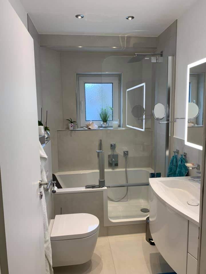 Jasna łazienka z wanną z niskim wejściem, kabiną prysznicową za szybą, umywalką z szafką i toaletą, oświetlona punktowo, z roślinami w oknie.