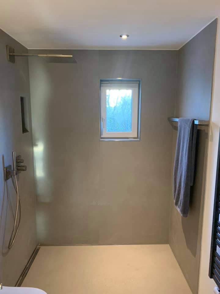 Minimalistyczna łazienka z szarą, gładką powierzchnią ścian, deszczownicą i małym oknem z widokiem na zieleń na zewnątrz.