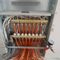 Rozdzielacz ogrzewania podłogowego z mosiężnymi zaworami i białymi pokrętłami, zamontowany w surowym otoczeniu z widocznymi przewodami i manometrem.