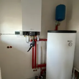 Biały piec grzewczy Viessmann zamontowany na ścianie, połączony z czerwonymi rurami i białym zbiornikiem wody z niebieskim naczyniem wzbiorczym. Na ścianie widoczny termostat. Na podłodze karton...