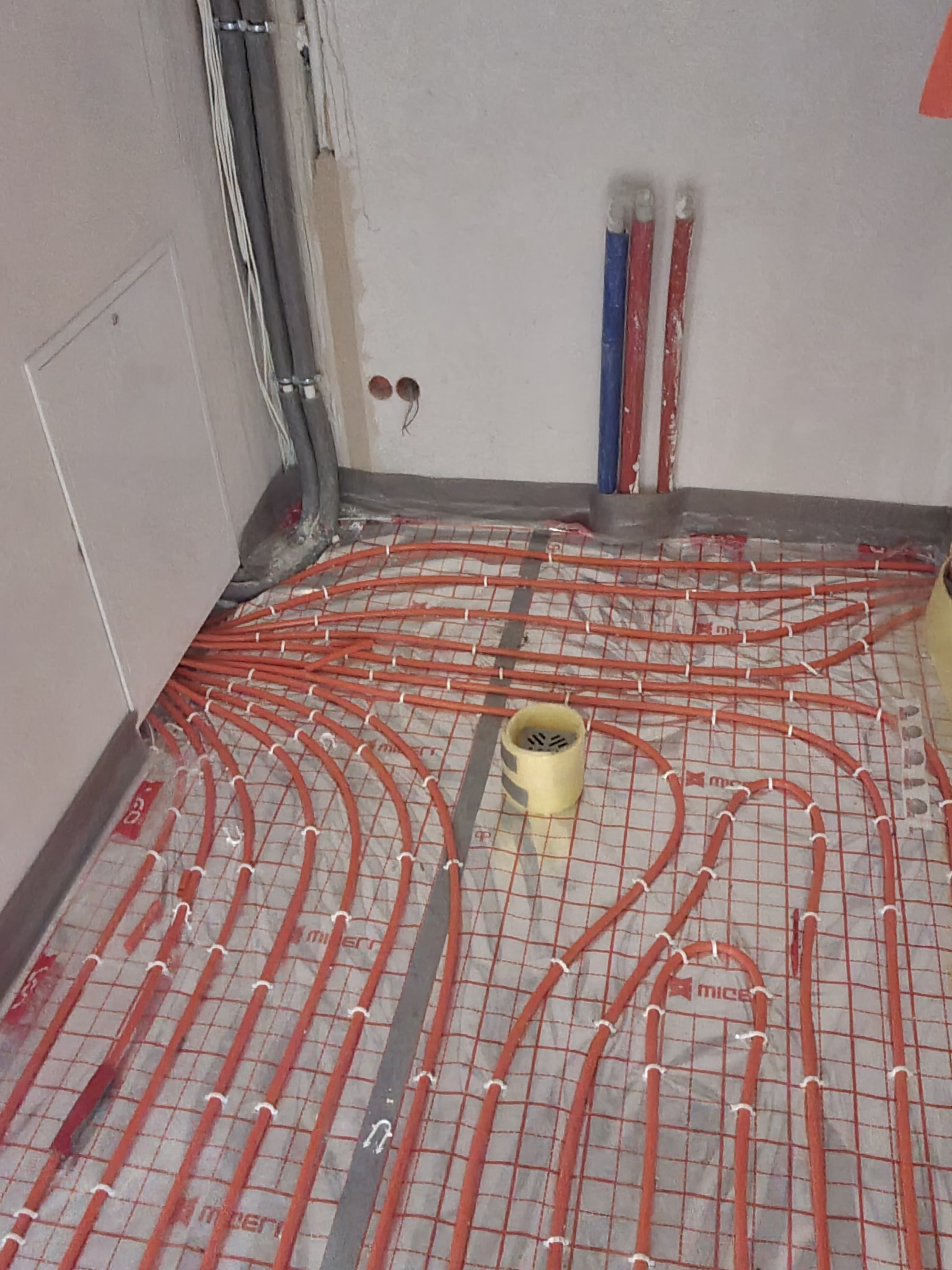 Pomarańczowe rury ogrzewania podłogowego ułożone na siatce, przymocowane białymi opaskami, widoczne fragmenty ścian i pionowe rury doprowadzające.
