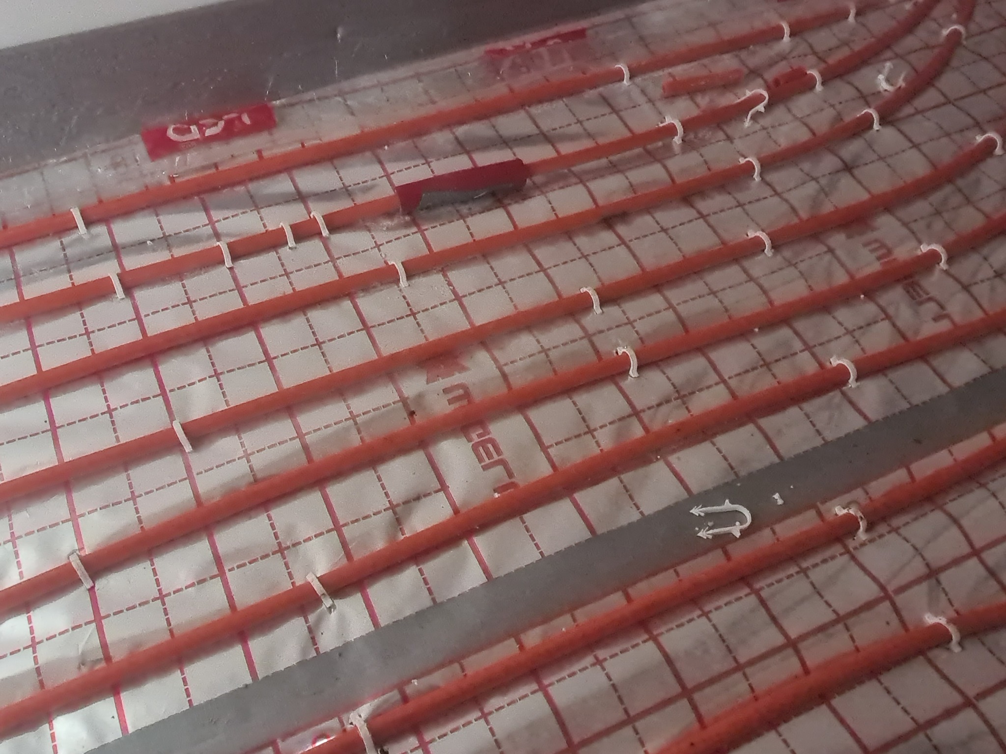 Pomarańczowe rury grzewcze przymocowane białymi uchwytami do folii z nadrukowaną siatką, tworzące system ogrzewania podłogowego.