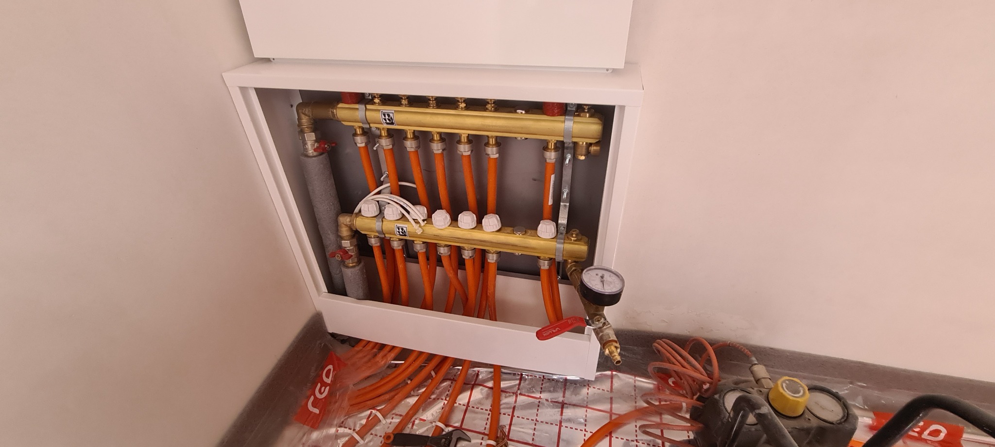 Rozdzielacz ogrzewania podłogowego w białej szafce, pomarańczowe rury, manometr i narzędzia widoczne na folii izolacyjnej.