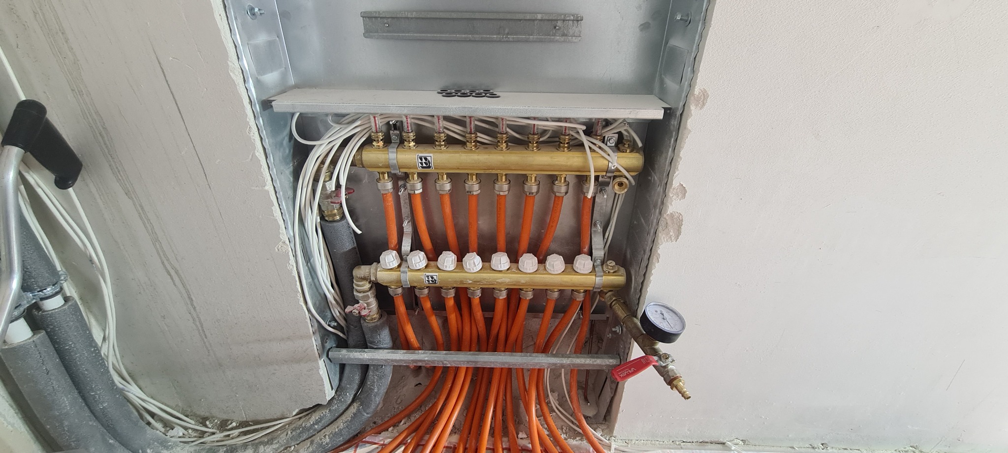 Rozdzielacz ogrzewania podłogowego z mosiężnymi zaworami i białymi pokrętłami, zamontowany w surowym otoczeniu z widocznymi przewodami i manometrem.