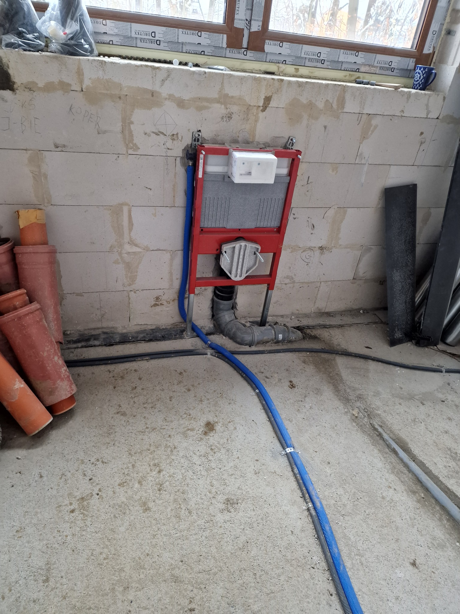 Stelaż podtynkowy WC zamontowany na surowej ścianie z betonu komórkowego, widoczne rury doprowadzające wodę i odpływ kanalizacyjny oraz fragmenty instalacji grzewczej.