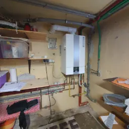 Kocioł gazowy kondensacyjny marki Wolf zamontowany na ścianie w piwnicy, widoczne rury instalacji grzewczej i gazowej, oraz półki z różnymi przedmiotami.