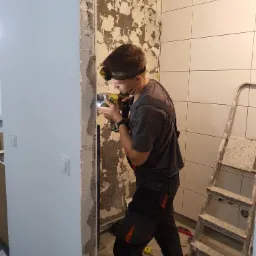 Pracownik w kombinezonie roboczym wierci w ścianie w łazience z częściowo położonymi białymi płytkami, przy użyciu wiertarki akumulatorowej z oświetleniem LED na głowie, obok stoi drabina malarska.