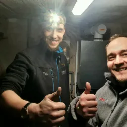 Dwóch uśmiechniętych monterów z kciukami w górę, jeden z czołową latarką, na tle kotła grzewczego w piwnicy z surowymi ścianami.