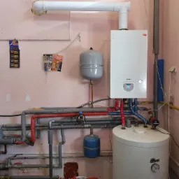 Kocioł gazowy marki Termet zawieszony na ścianie, z podłączonymi rurami w kolorach czerwonym i niebieskim, zbiornik wyrównawczy oraz bojler Kospel z widocznym manometrem, całość w pomieszczeniu...