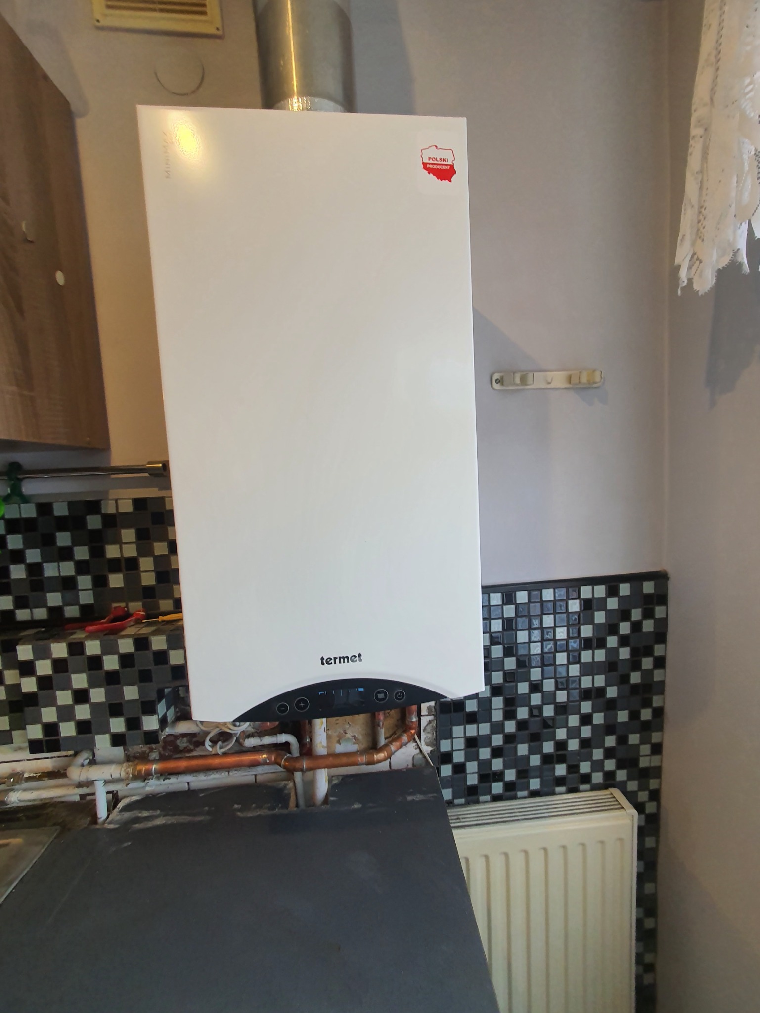 Biały piec gazowy marki Termet z naklejką 'Polski Producent' zamontowany na ścianie z mozaiką, widoczne rury miedziane i fragment grzejnika.