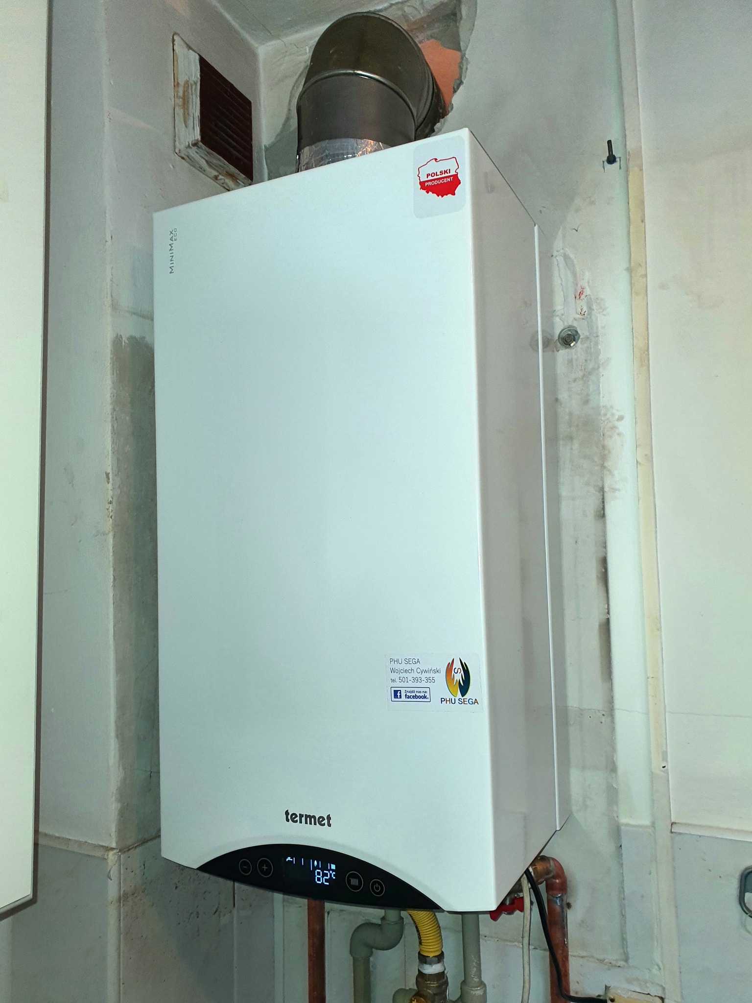 Biały piec gazowy Termet Minimax Eco zawieszony na ścianie z widocznymi przyłączami, naklejką 'Polski Producent' i panelem sterowania wskazującym temperaturę 82°C. Na ścianie widoczne ślady montażu...