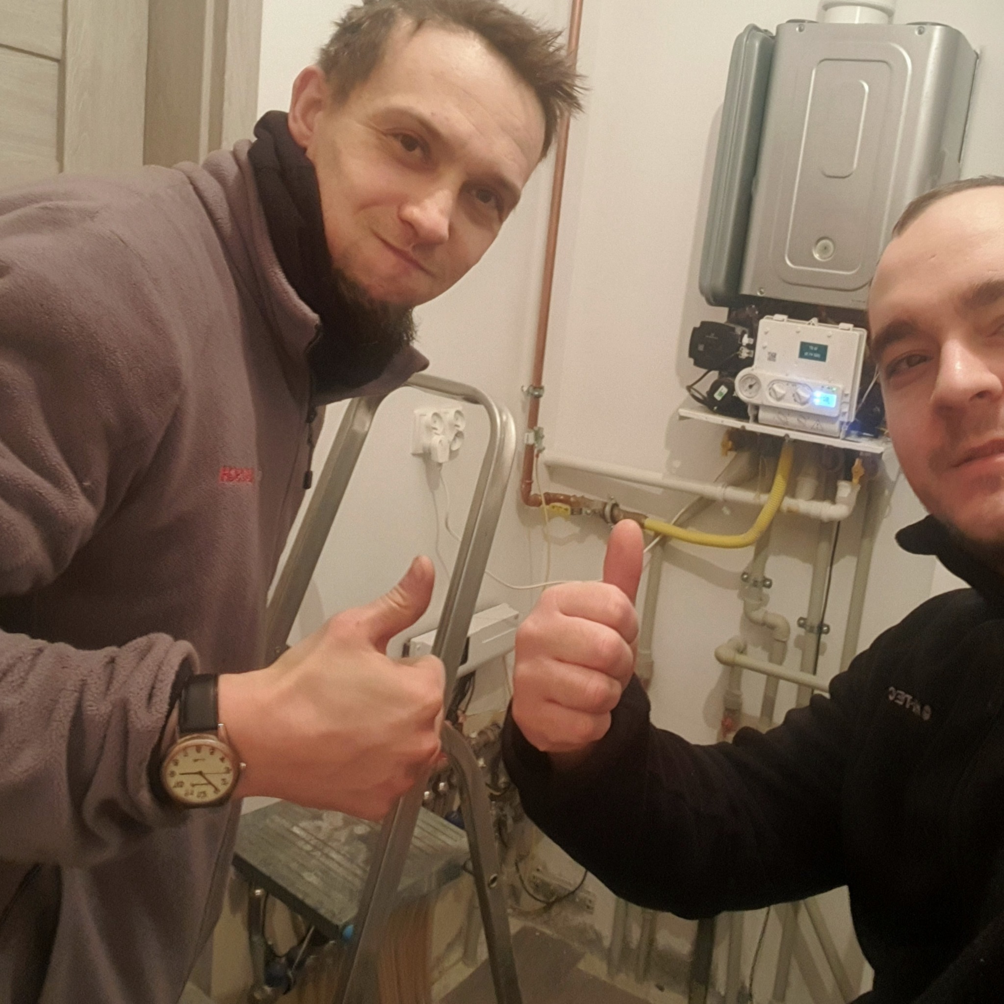 Dwóch instalatorów z podniesionymi kciukami na tle zamontowanego pieca gazowego, widoczna drabina i instalacja gazowa.