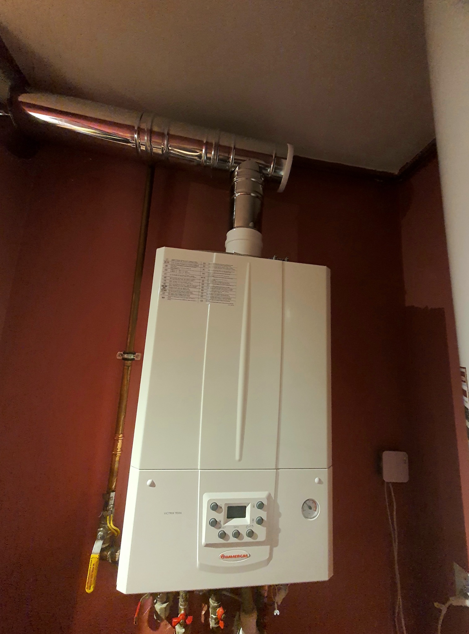 Biały piec gazowy marki Immergas zamontowany na ścianie w pomieszczeniu z brązową ścianą i widocznymi rurami, z chromowanym kominem odprowadzającym spaliny.